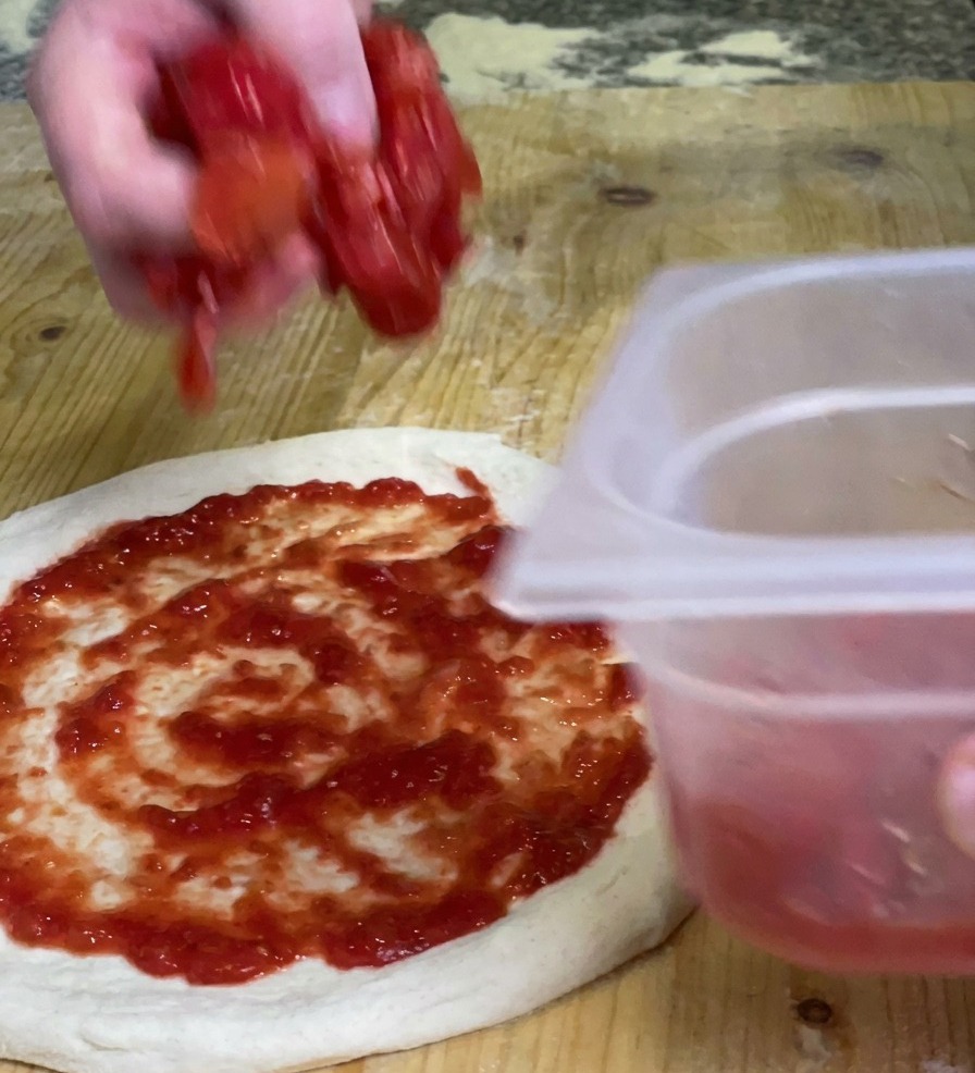 Dłoń nakładająca kawałki gęstego, zgniecionego miąższu pomidorowego na spód pizzy pokryty gładkim czerwonym sosem.
