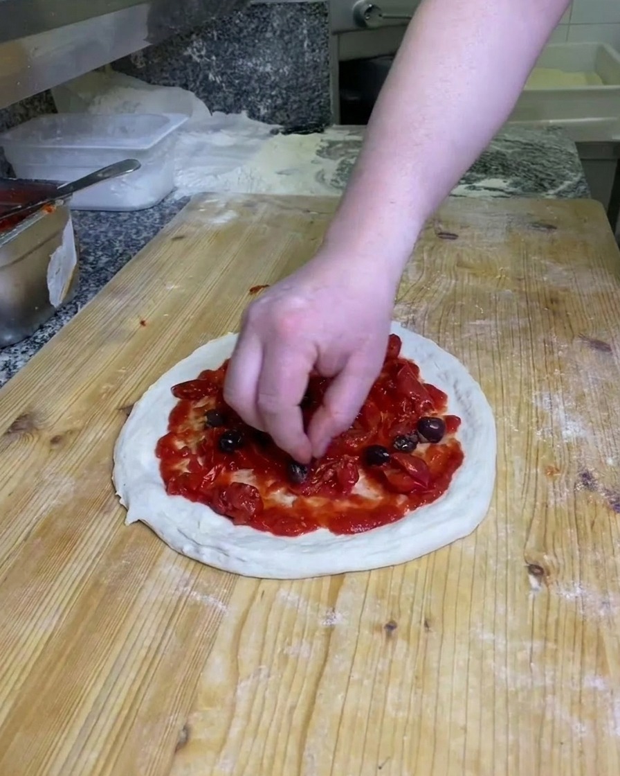 Dłoń układająca ciemne, całe oliwki na cieście do pizzy posmarowanym pomidorami, na oprószonej mąką drewnianej powierzchni.