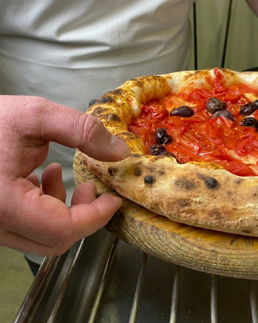 Palec kucharza naciskający na miękki, przypieczony i puszysty brzeg świeżo upieczonej pizzy Marinara.