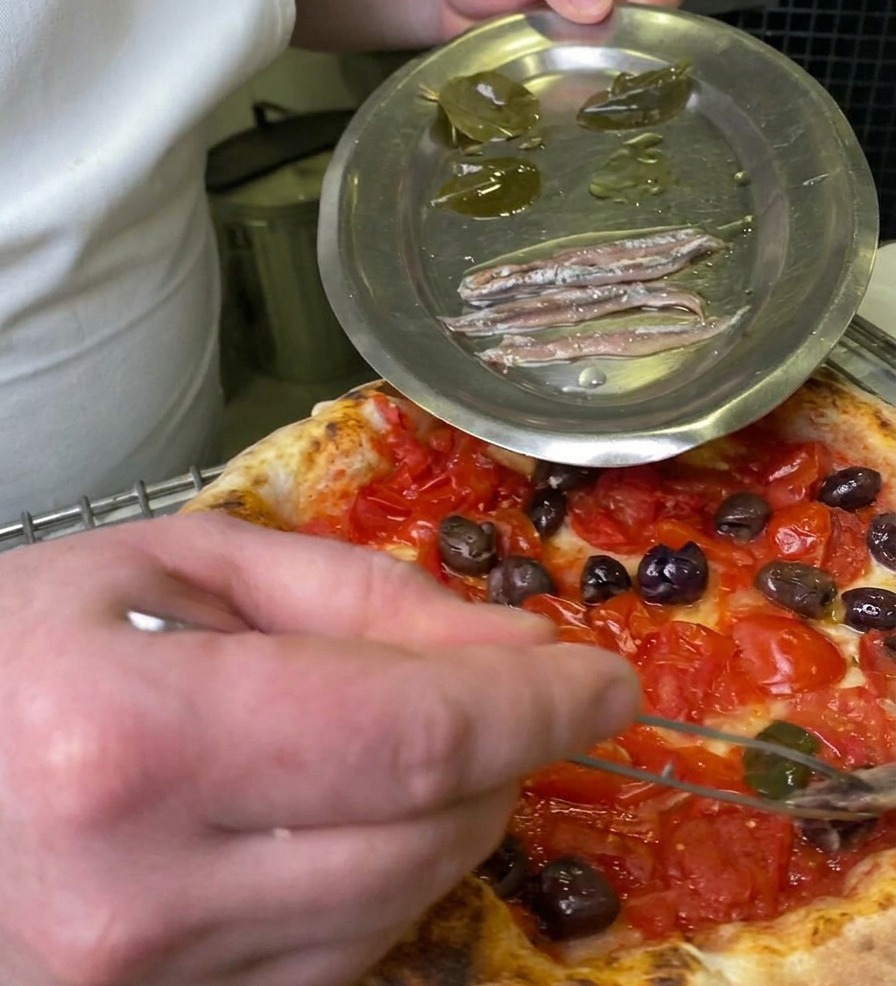 Kucharz używający pęsety do chwytania marynowanego liścia laurowego z metalowego talerza, aby położyć go na pizzy z pomidorami i oliwkami.