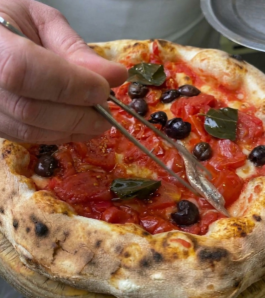 Zbliżenie na srebrzysty filet anchois układany na cieście do pizzy za pomocą metalowej pęsety kulinarnej.