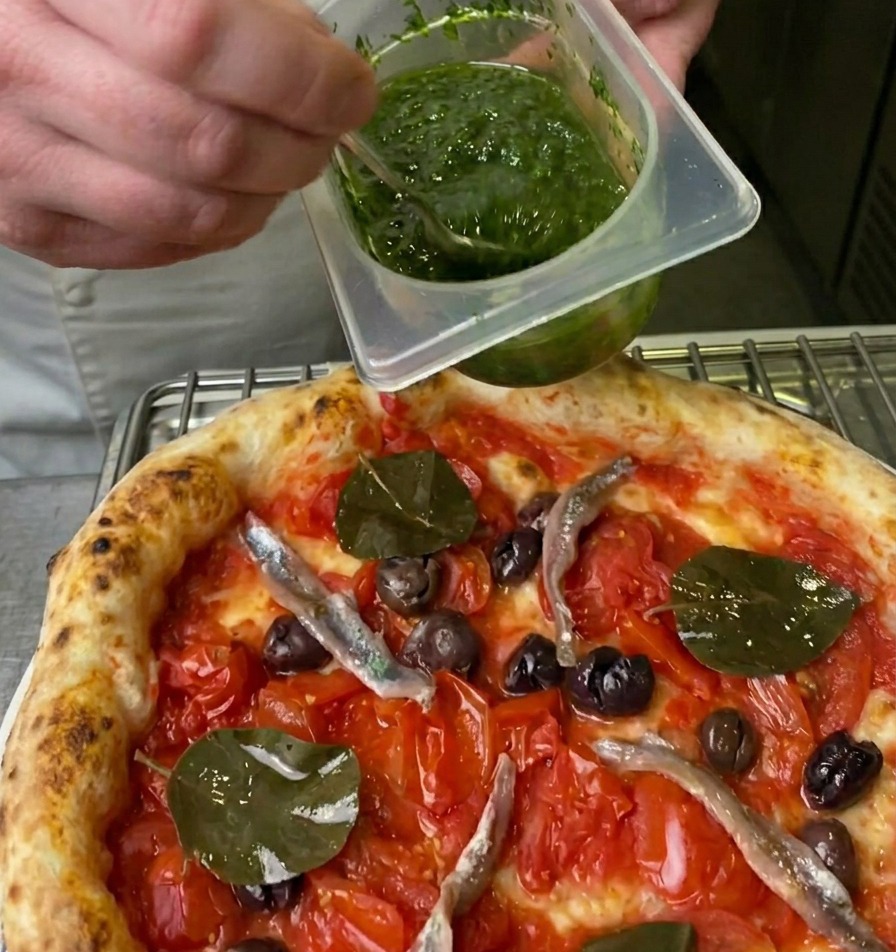Kucharz skrapiający pizzę jaskrawozielonym sosem ziołowym z plastikowego pojemnika przy użyciu małej łyżeczki.