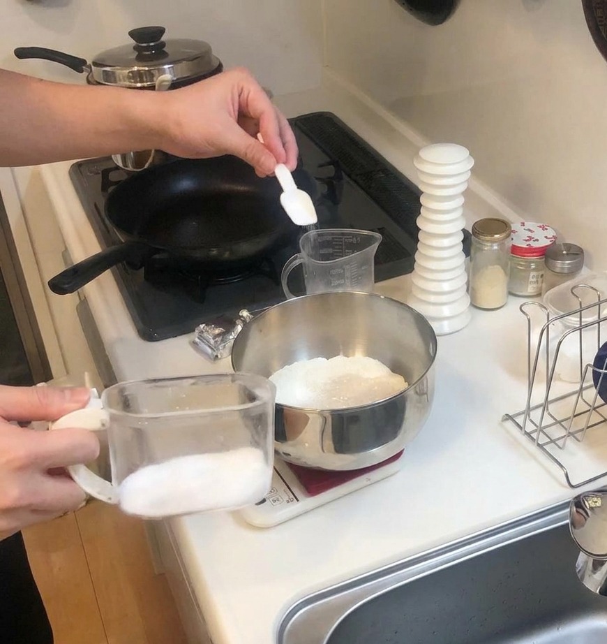 Mała biała miarka dodająca cukier do miski z mąką stojącej na wadze kuchennej.