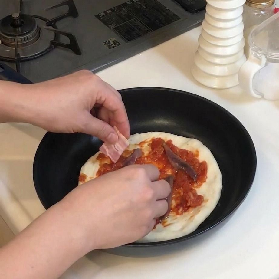 Dłonie kładące kawałki surowego boczku na pizzę z sosem i anchois.