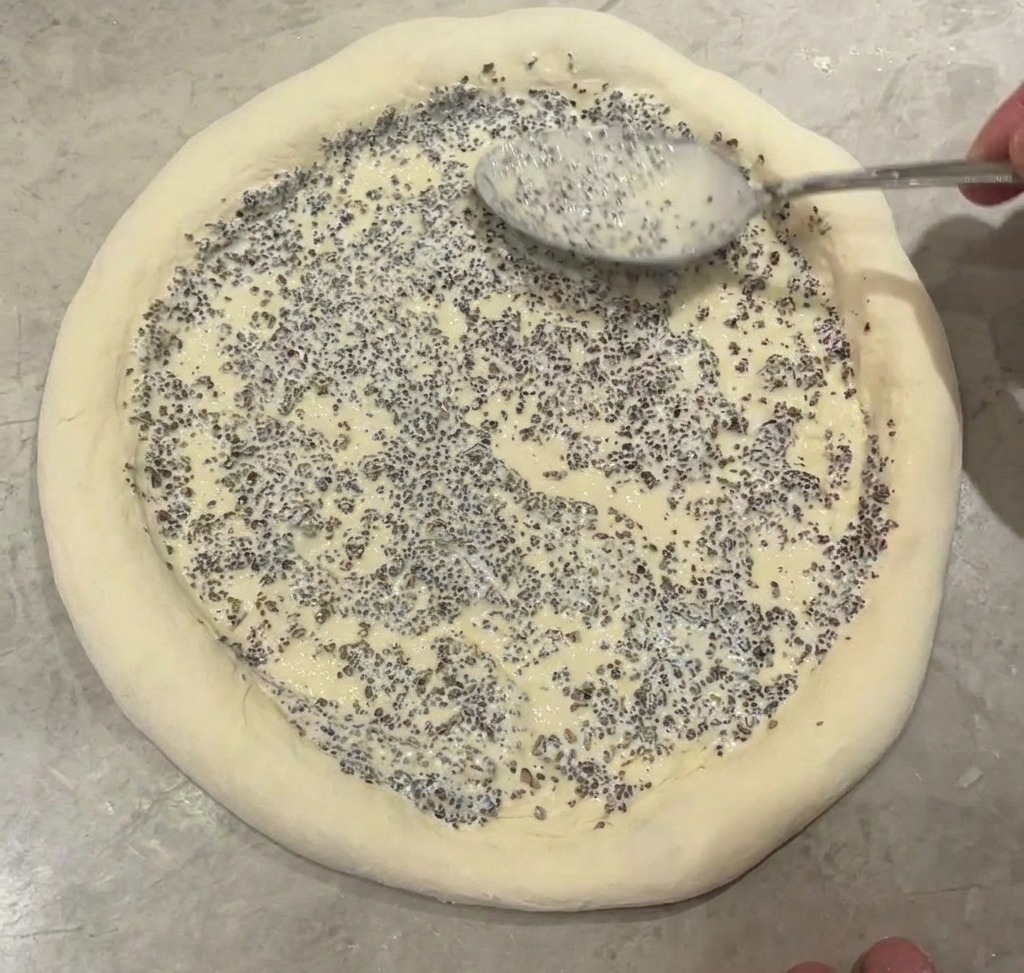 Metalowa łyżka rozprowadzająca gęsty sos truflowy z widocznymi drobinkami na środku bazy pizzy.