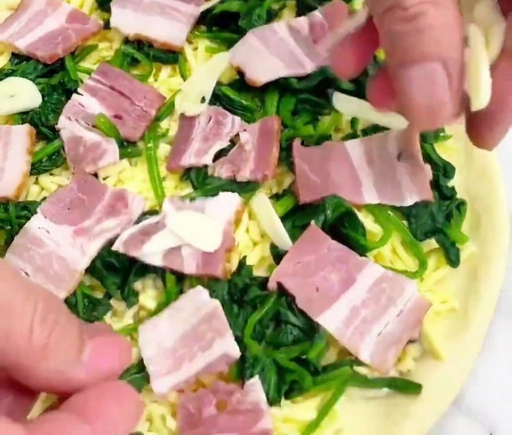 Dłoń sypiąca cienkie, przezroczyste plasterki czosnku na surową pizzę ze szpinakiem, boczkiem i serem.
