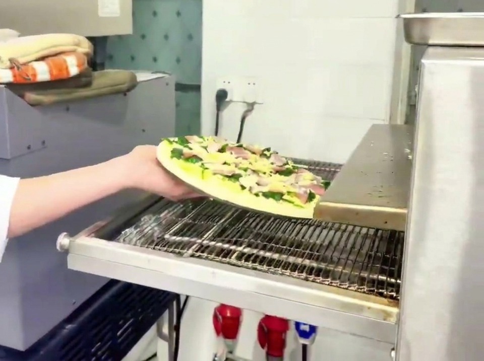 Pracownik kładący surową pizzę ze szpinakiem i boczkiem na metalowy pas transmisyjny profesjonalnego pieca.