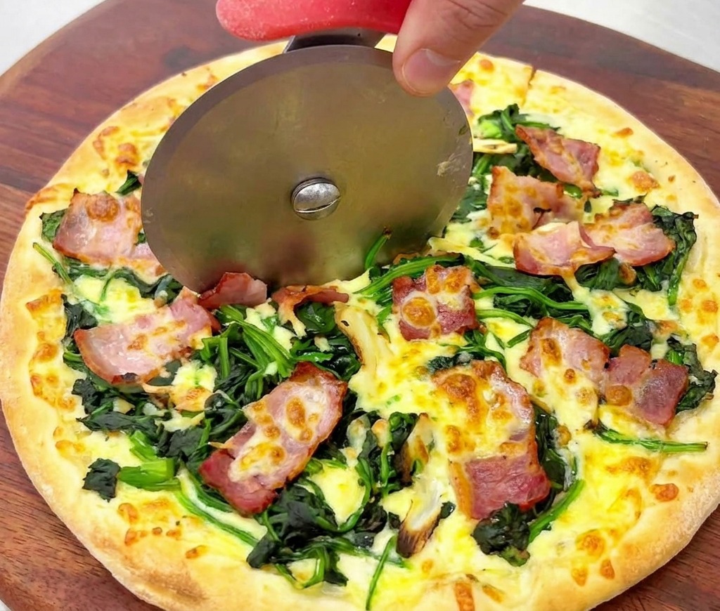 Zbliżenie na nóż krążkowy przecinający złocistą pizzę z upieczonym szpinakiem i chrupiącym boczkiem.