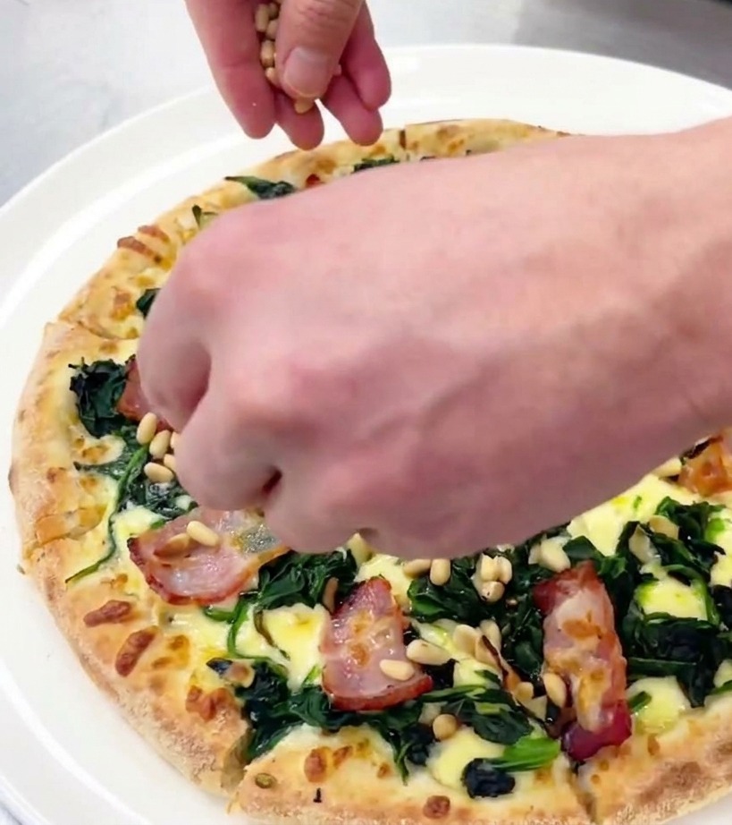Dłoń rozsypująca małe prażone orzeszki piniowe na pokrojonej pizzy leżącej na białym talerzu.