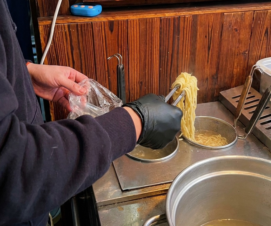Osoba w czarnych rękawiczkach używająca szczypiec do wyciągania długiego, żółtego makaronu ramen z garnka z gotującą się wodą.