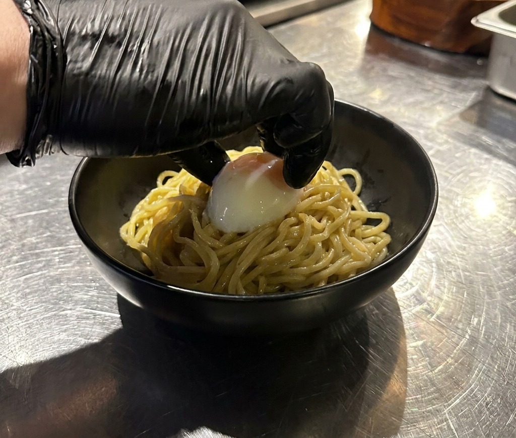 Dłoń w czarnej rękawiczce kładzie całe jajko na miękko na porcji makaronu ramen w czarnej misce.