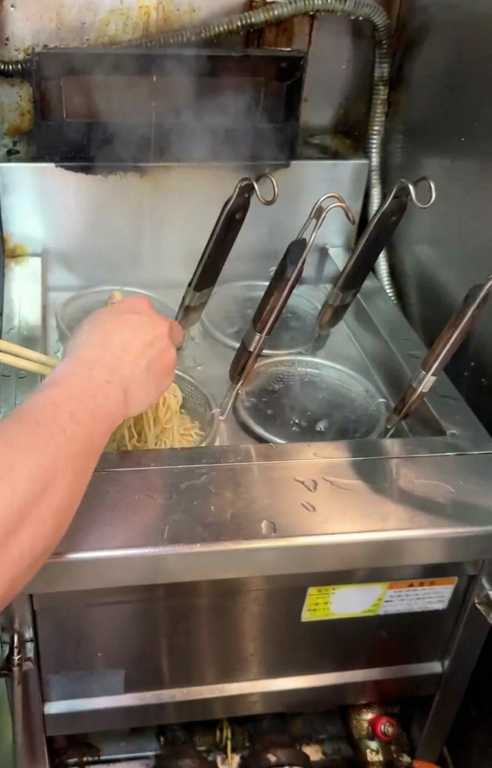 Kucharz wyjmujący sitko z makaronem z wrzątku i energicznie nim potrząsający, aby odcedzić ramen.