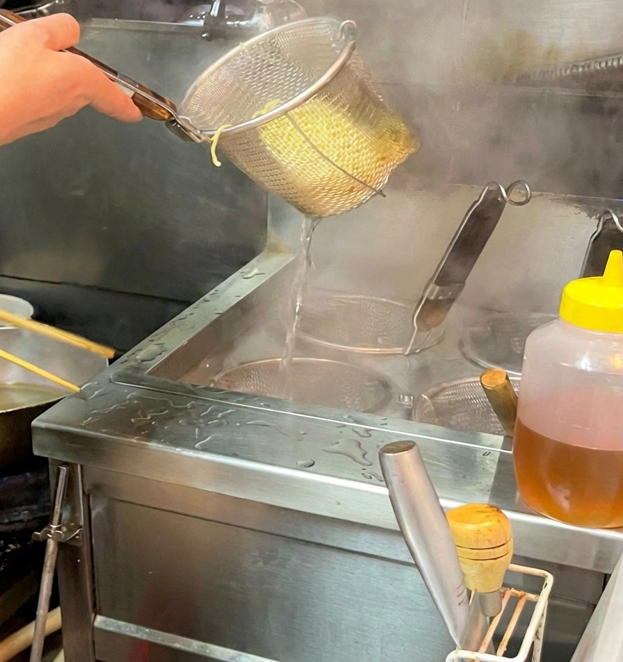 Dłoń trzymająca sitko nad miską, przesypująca ugotowany makaron ramen do parującego, ciemnego wywaru.