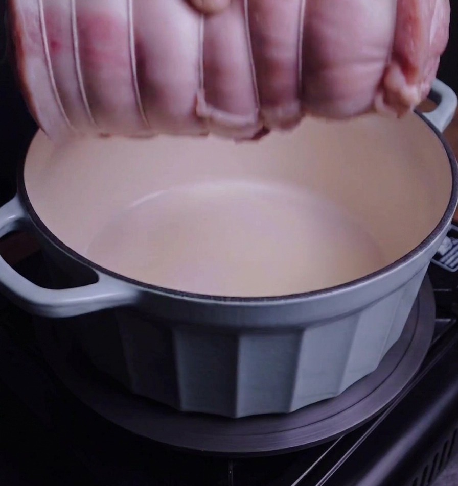Związany rulon boczku wkładany ręką do błękitnego garnka żeliwnego typu Dutch oven.