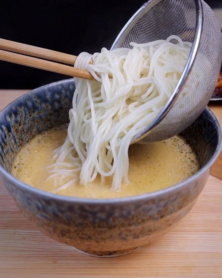 Sitko ze stali nierdzewnej przekładające długi biały makaron do miski z żółtym wywarem ramen.