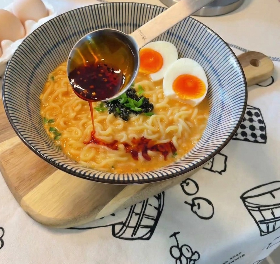Czerwony olej chili wylewany z metalowej miarki na gotową miskę ramenu z jajkami, cebulą i wodorostami.