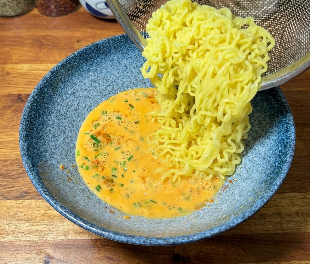 Ugotowany makaron ramen przekładany z sitka do niebieskiej miski z pomarańczowym, kremowym bulionem.