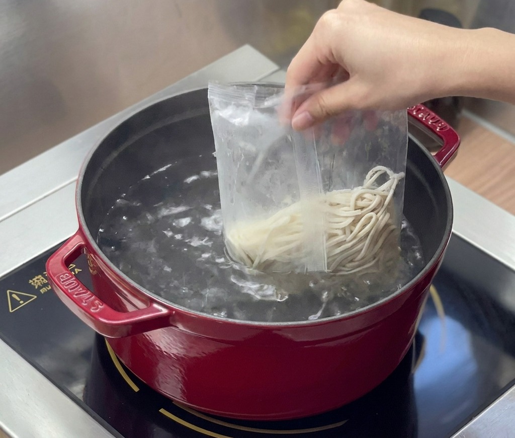 Świeży makaron ramen wsypywany z foliowego woreczka do czerwonego garnka z wrzącą wodą.
