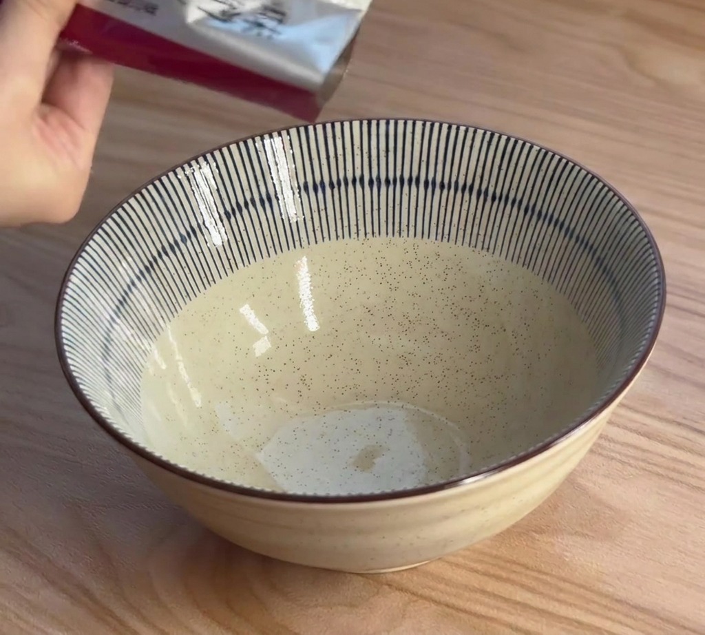 Dłoń wyciskająca gęstą pastę miso z saszetki do ceramicznej miski w niebiesko-białe pasy.