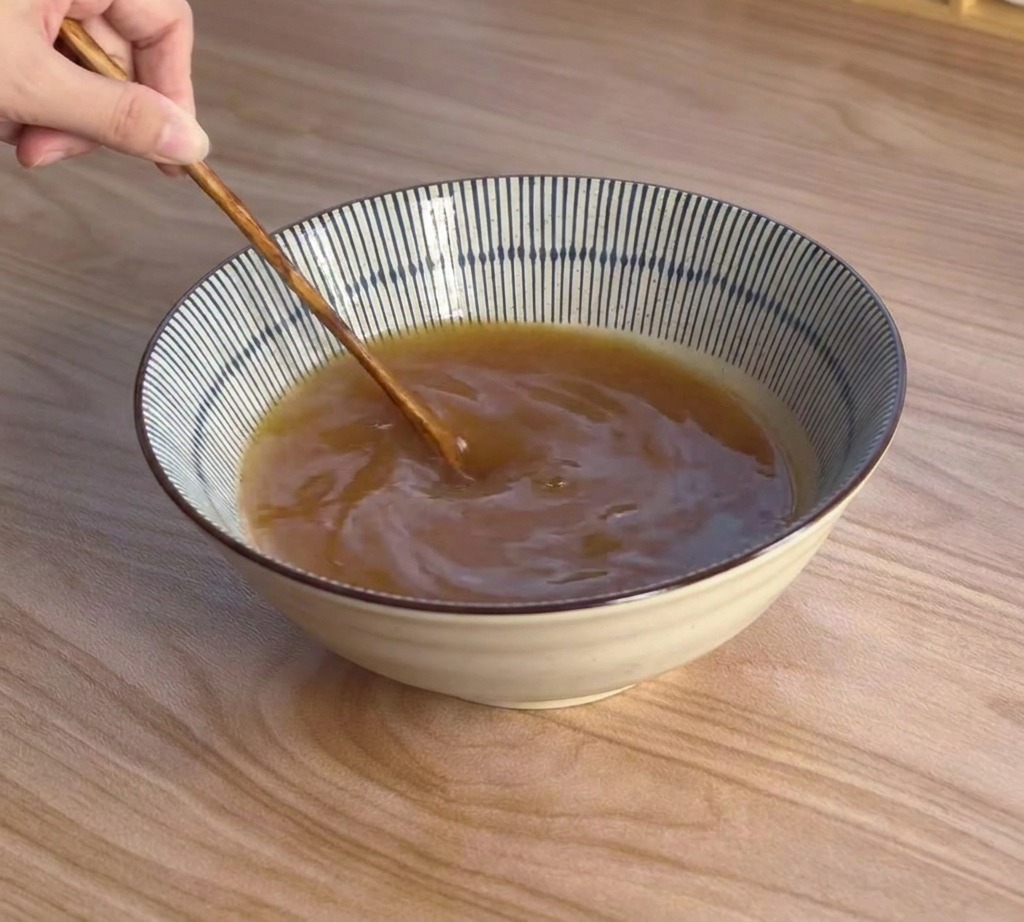 Dłoń używająca drewnianej łyżki do mieszania brązowego bulionu miso w ceramicznej misce.