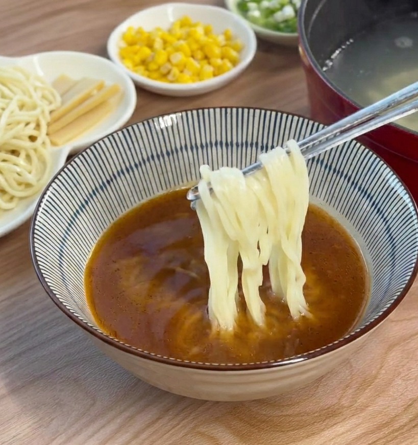 Ugotowany makaron ramen dodawany pałeczkami do miski z brązowym bulionem miso.