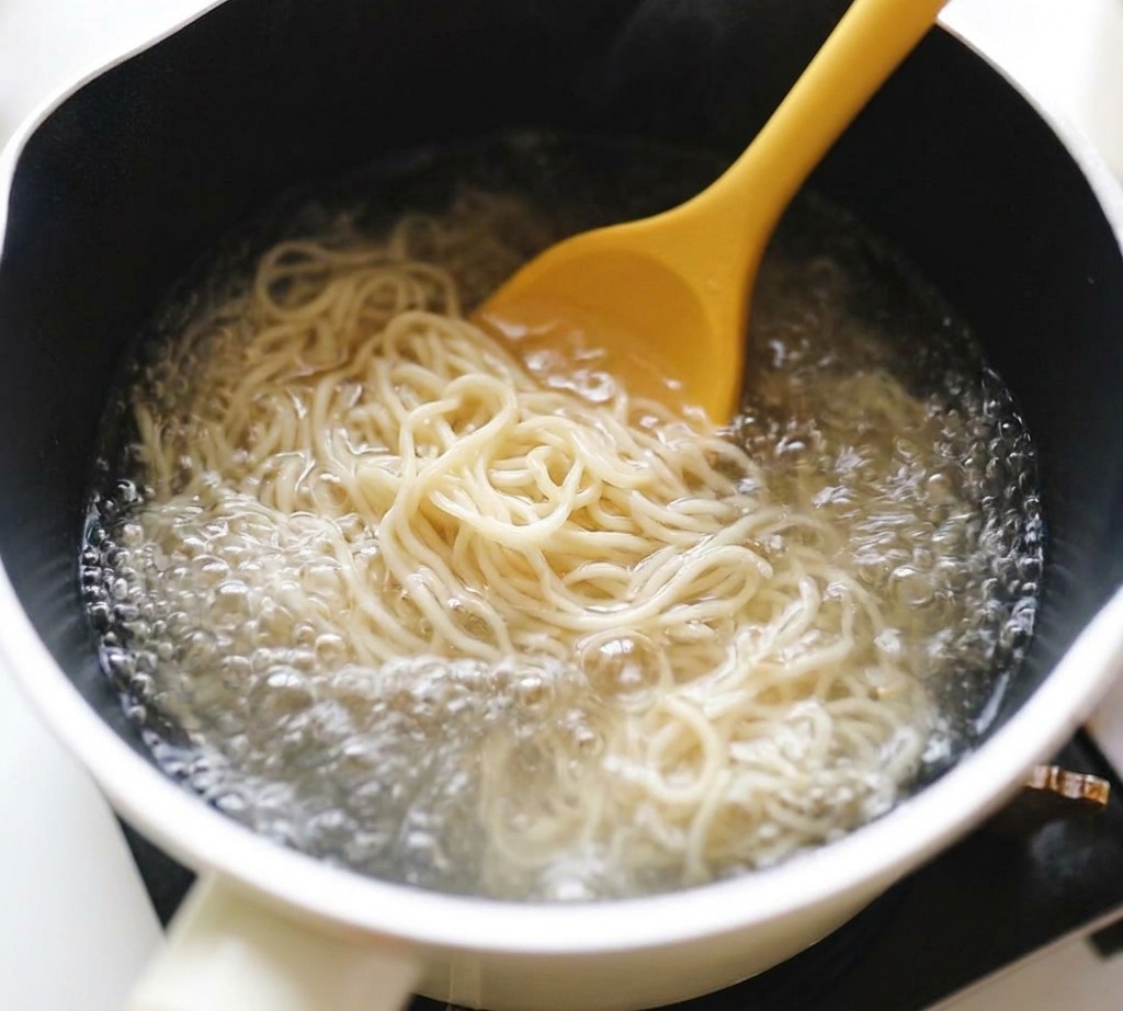 Falisty makaron ramen mieszany żółtą łyżką w garnku z mocno gotującą się wodą.