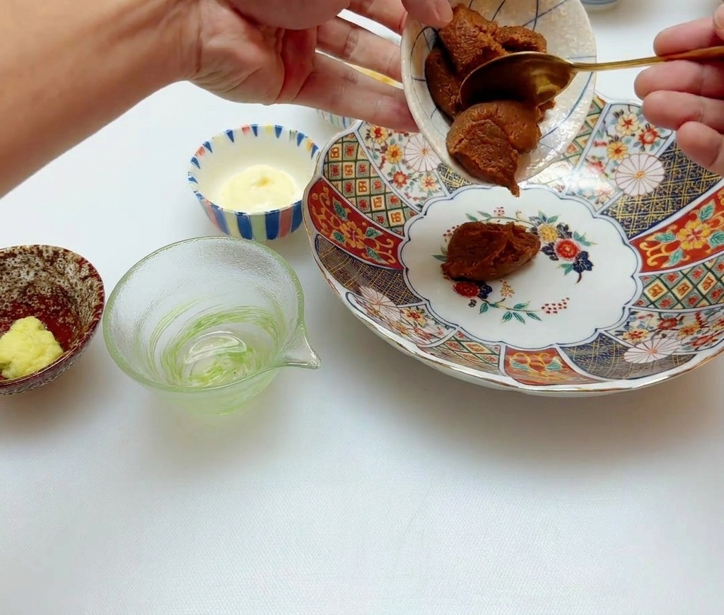 Dłoń nakładająca gęstą brązową pastę miso ze złotą łyżką do dużej, wzorzystej miski ceramicznej.