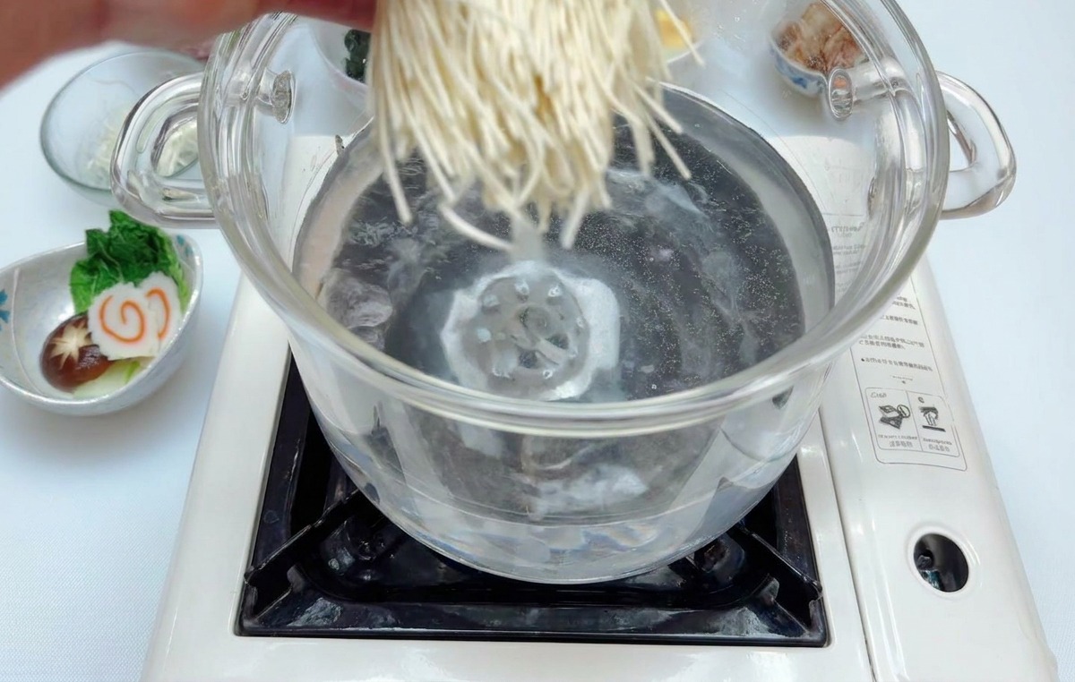 Dłoń wrzucająca porcję białego makaronu ramen do szklanego garnka z wrzącą wodą na przenośnej płycie indukcyjnej.