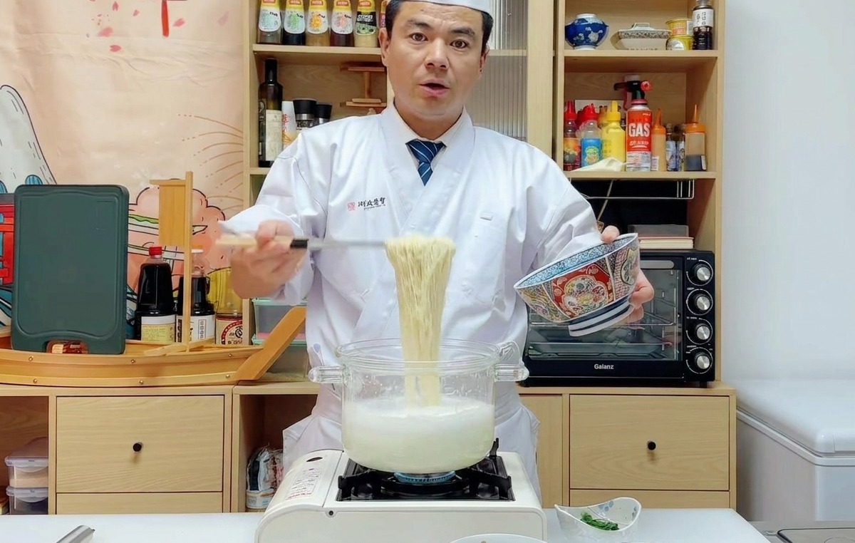 Szef kuchni Wu podnoszący sitkiem dużą porcję ugotowanego makaronu ramen i przekładający go do niebieskiej, wzorzystej miski.