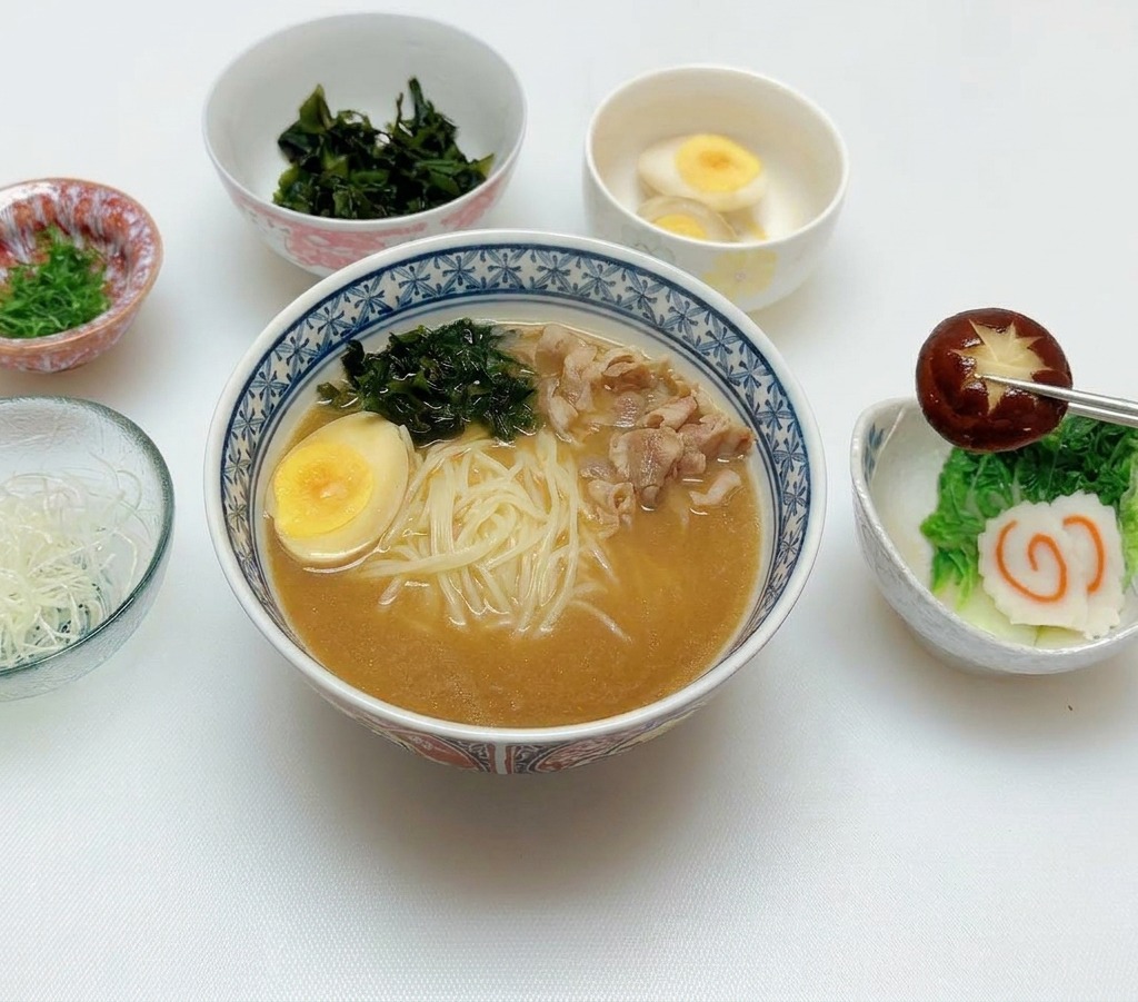 Pałeczki kładące ciemnobrązowy, nacięty grzyb shiitake na dodatki w misce miso ramen, obok jajka i ciastek rybnych.