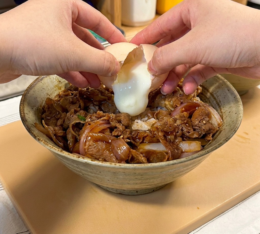 Zbliżenie na dłonie rozbijające miękkie jajko onsen nad miską z wołowiną teriyaki i ryżem.