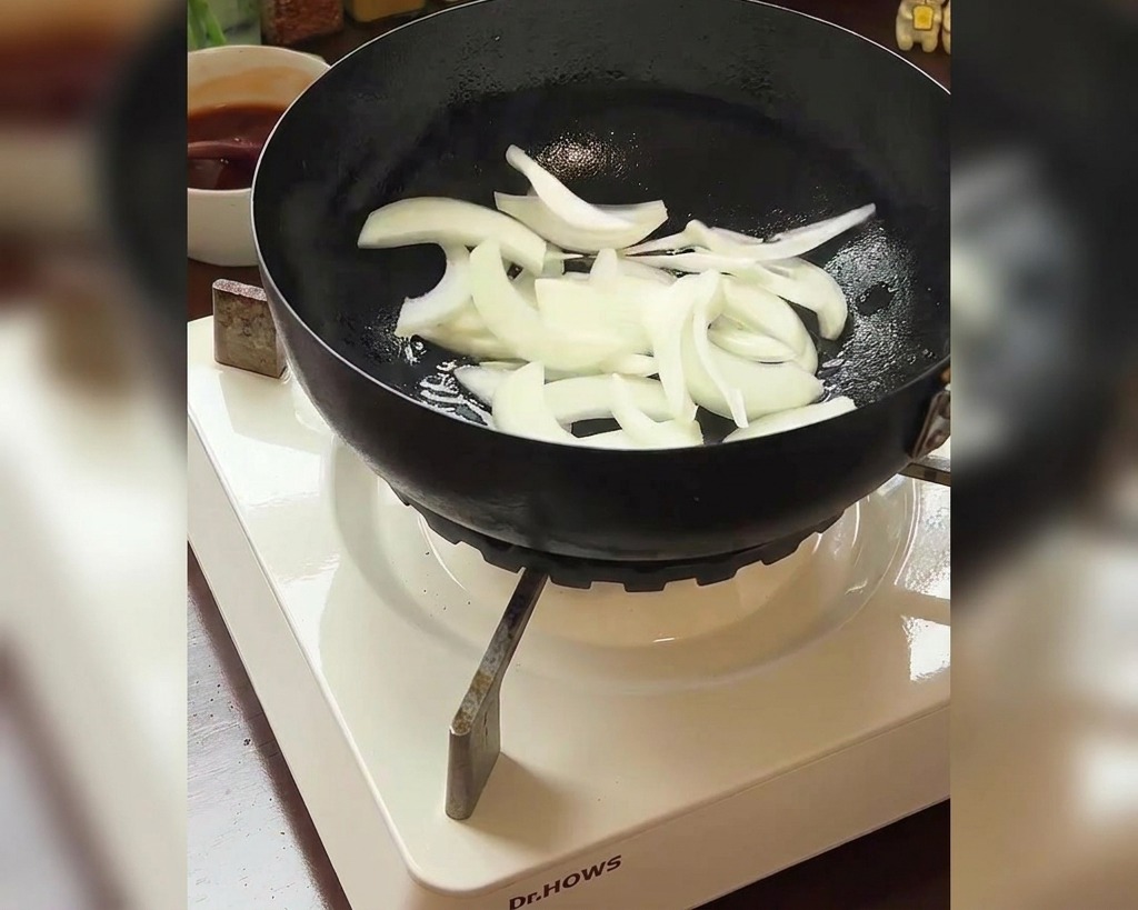Pokrojona cebula dodawana na patelnię, w tle widoczna miska czerwonego kimchi.