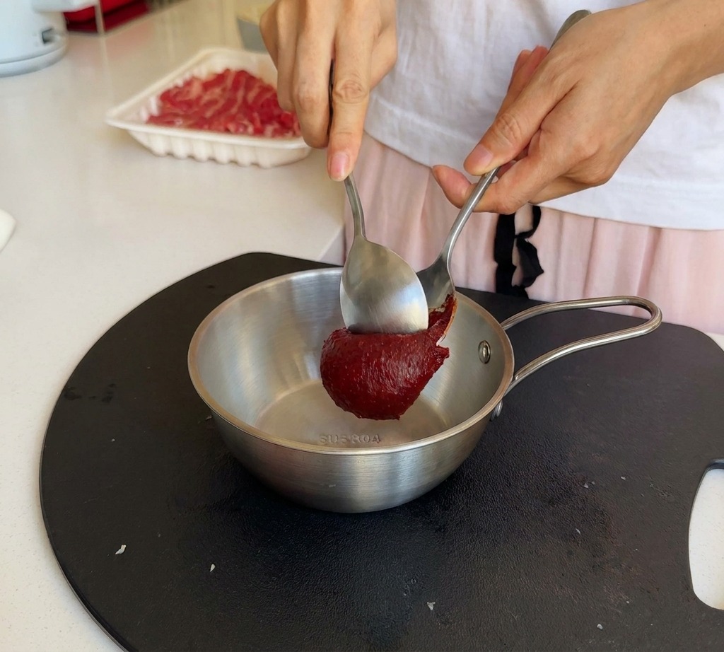Osoba dodająca dużą łyżkę gęstej, czerwonej koreańskiej pasty chili do metalowej miseczki.