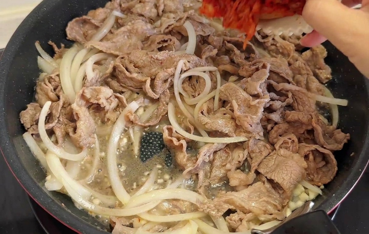 Ręka wlewająca z białej miseczki posiekane kimchi i zieloną paprykę na patelnię z wołowiną i cebulą.