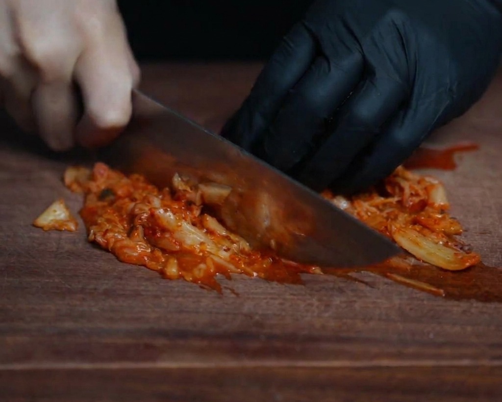 Zbliżenie szefa kuchni drobno siekającego jasnoczerwone kimchi na ciemnej drewnianej desce ostrym nożem.
