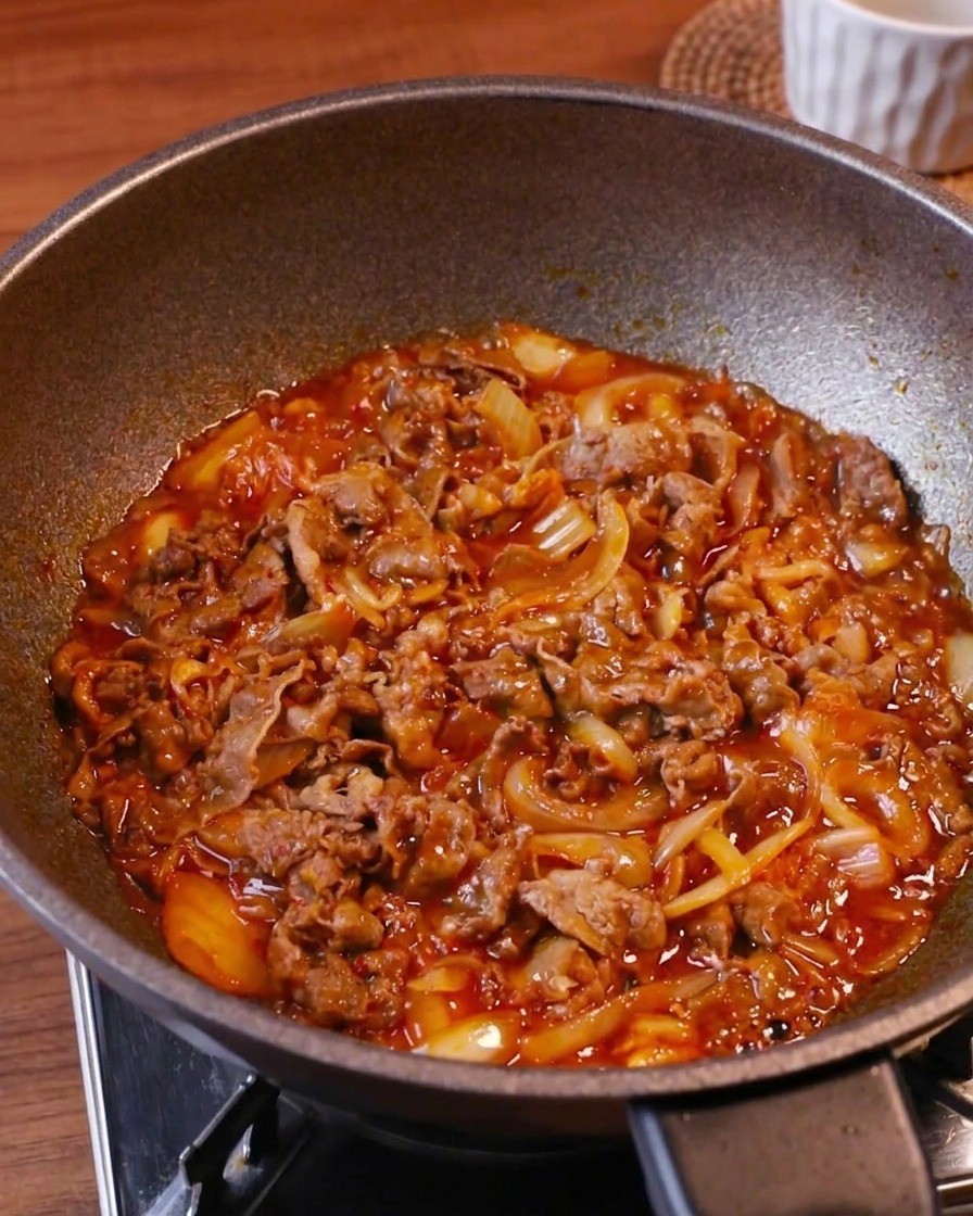 Wołowina, kimchi i cebula gotujące się w gęstym, bulgoczącym czerwonym sosie w woku.