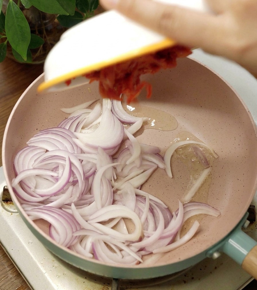 Różowa patelnia ceramiczna z pokrojoną czerwoną cebulą i porcją pikantnego kimchi dodawaną z białej miski.