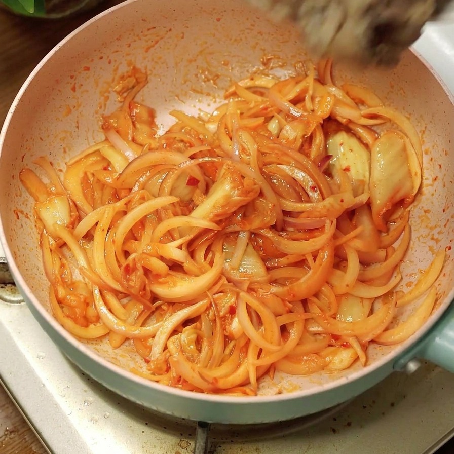 Ugotowana wołowina przesypywana z białej miski na różową patelnię z podsmażoną cebulą i kimchi.