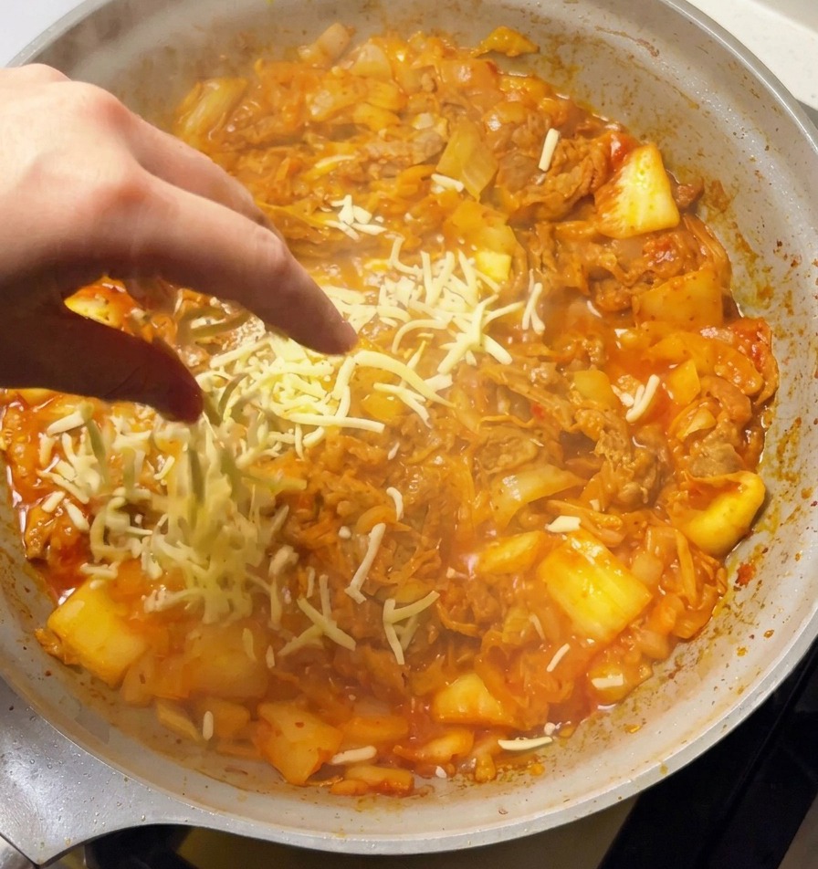 Dłoń posypująca obficie tartym białym serem mozzarella bulgoczące kimchi z wołowiną na patelni.