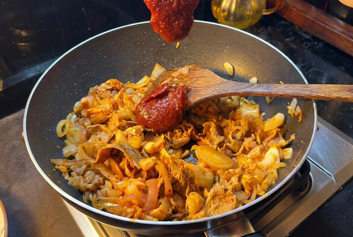Drewniana łopatka z dużą porcją gęstej, czerwonej koreańskiej pasty chili nad patelnią ze smażoną wołowiną i kimchi.