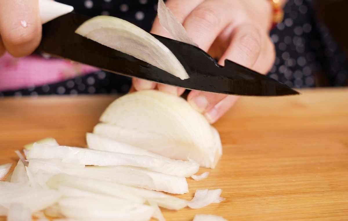 Osoba krojąca białą cebulę w cienkie paski na jasnej drewnianej desce kuchennej.