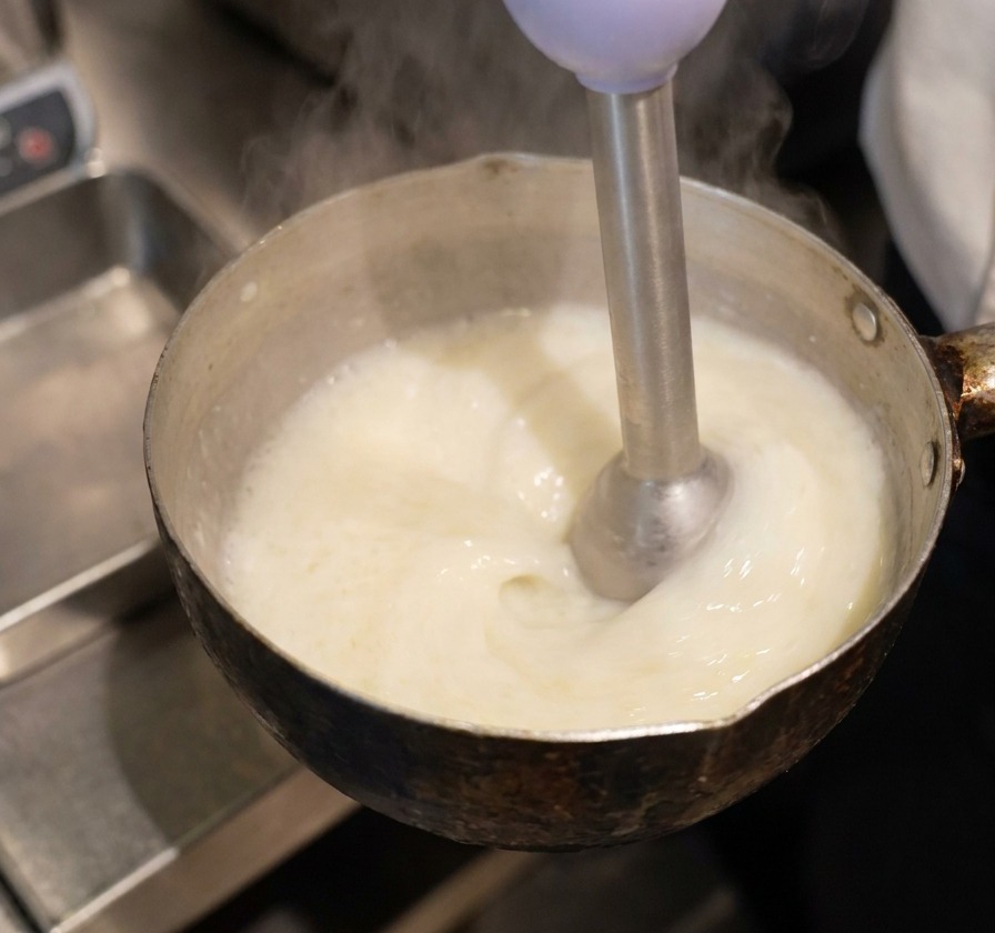 Blender ręczny zanurzony w garnku z płynem, tworzący gęstą białą pianę na powierzchni.