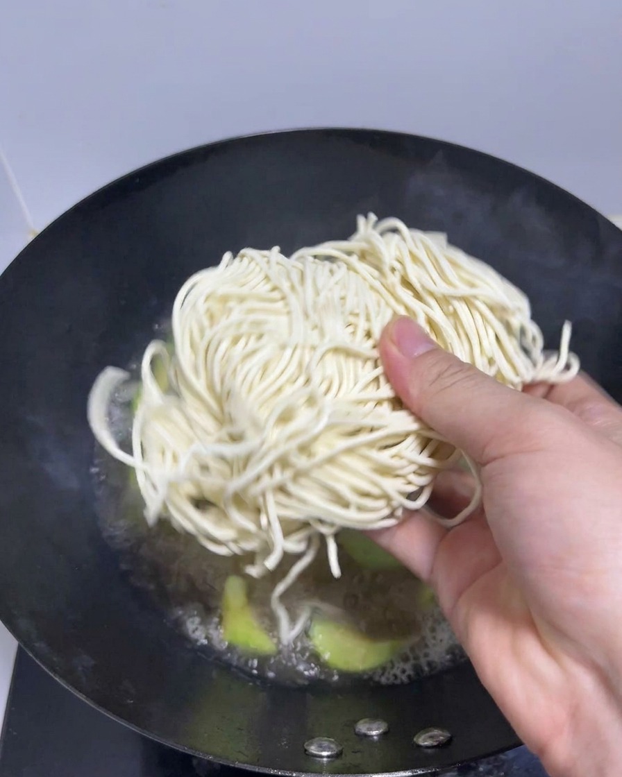 Dłoń wkładająca porcję świeżego makaronu ramen do woka z wrzącym wywarem.