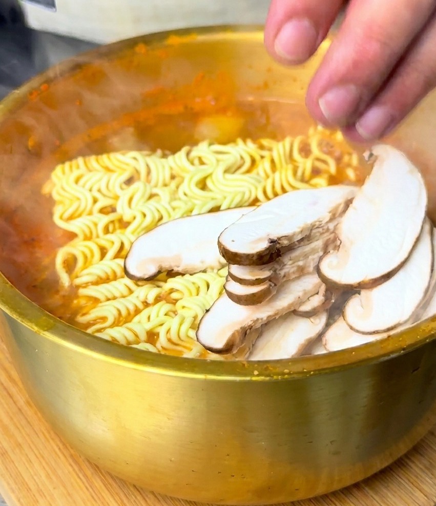 Dłoń kładąca grzyby shiitake na makaronie zanurzonym w gotującej się czerwonej zupie.