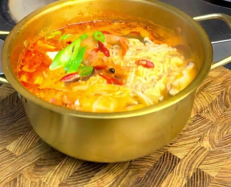 Pikantny ramen z owocami morza w złotym garnku, udekorowany plasterkami chili, na drewnianej desce.