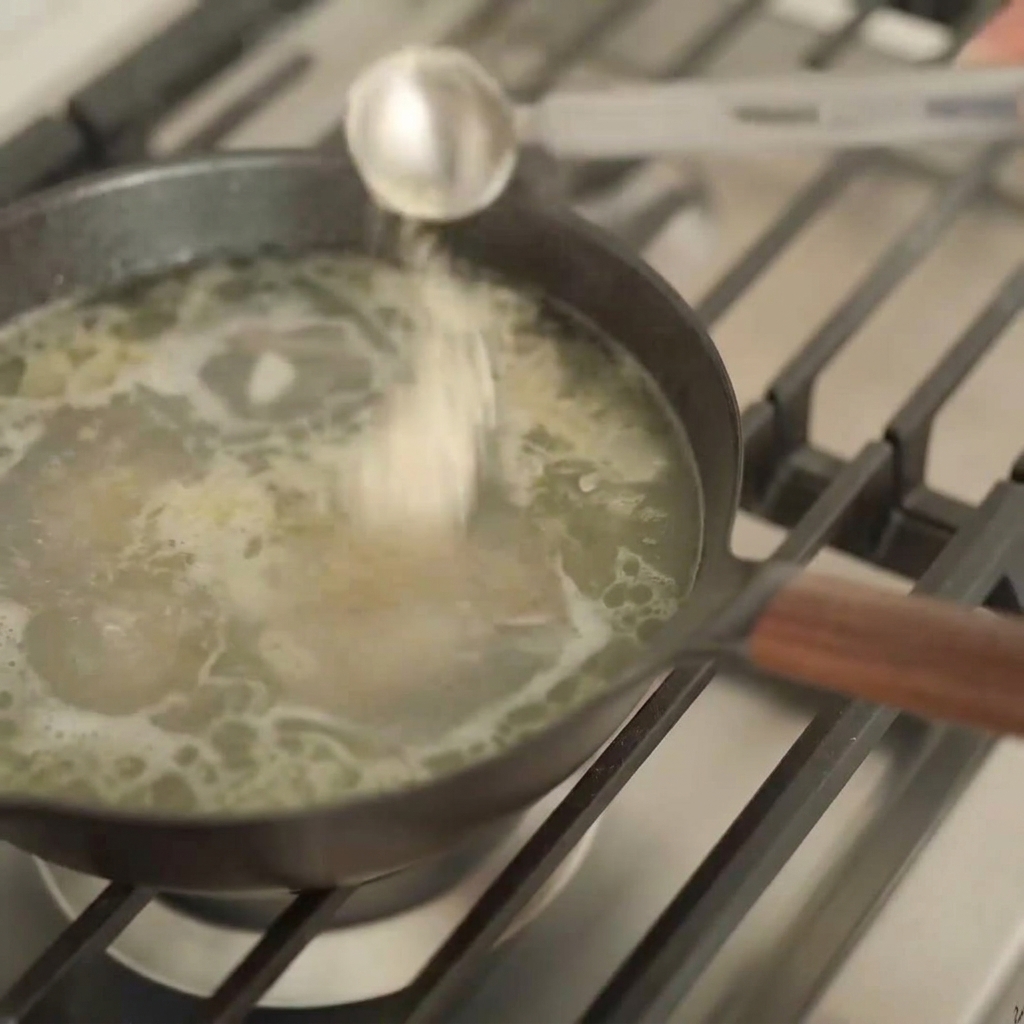 Srebrna łyżka miarowa wlewająca ciemny płyn do garnka z jasnym, bąbelkującym wywarem ramen.