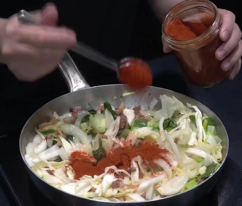 Jaskrawoczerwona papryka chili w proszku nakładana łyżką ze szklanego słoika na smażone warzywa i mięso.