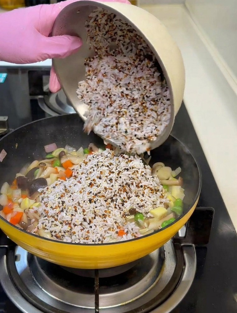 Ręka w różowej rękawiczce wsypująca mieszankę namoczonego ryżu i quinoa na patelnię z warzywami i grzybami.