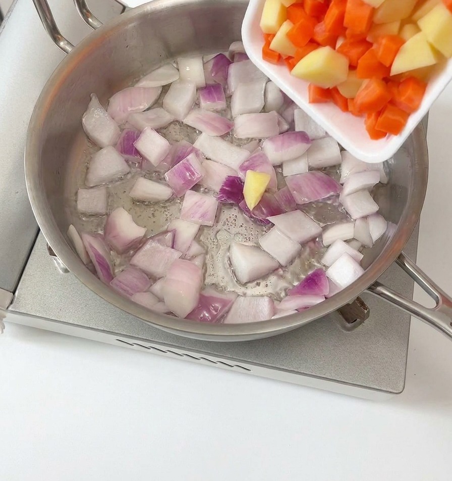 Pokrojone w kostkę ziemniaki i marchewka wsypywane na patelnię do podsmażonej czerwonej cebuli.