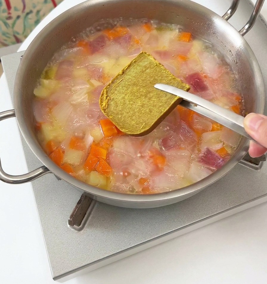 Metalowe szczypce trzymające brązową kostkę curry nad parującą patelnią z warzywami.